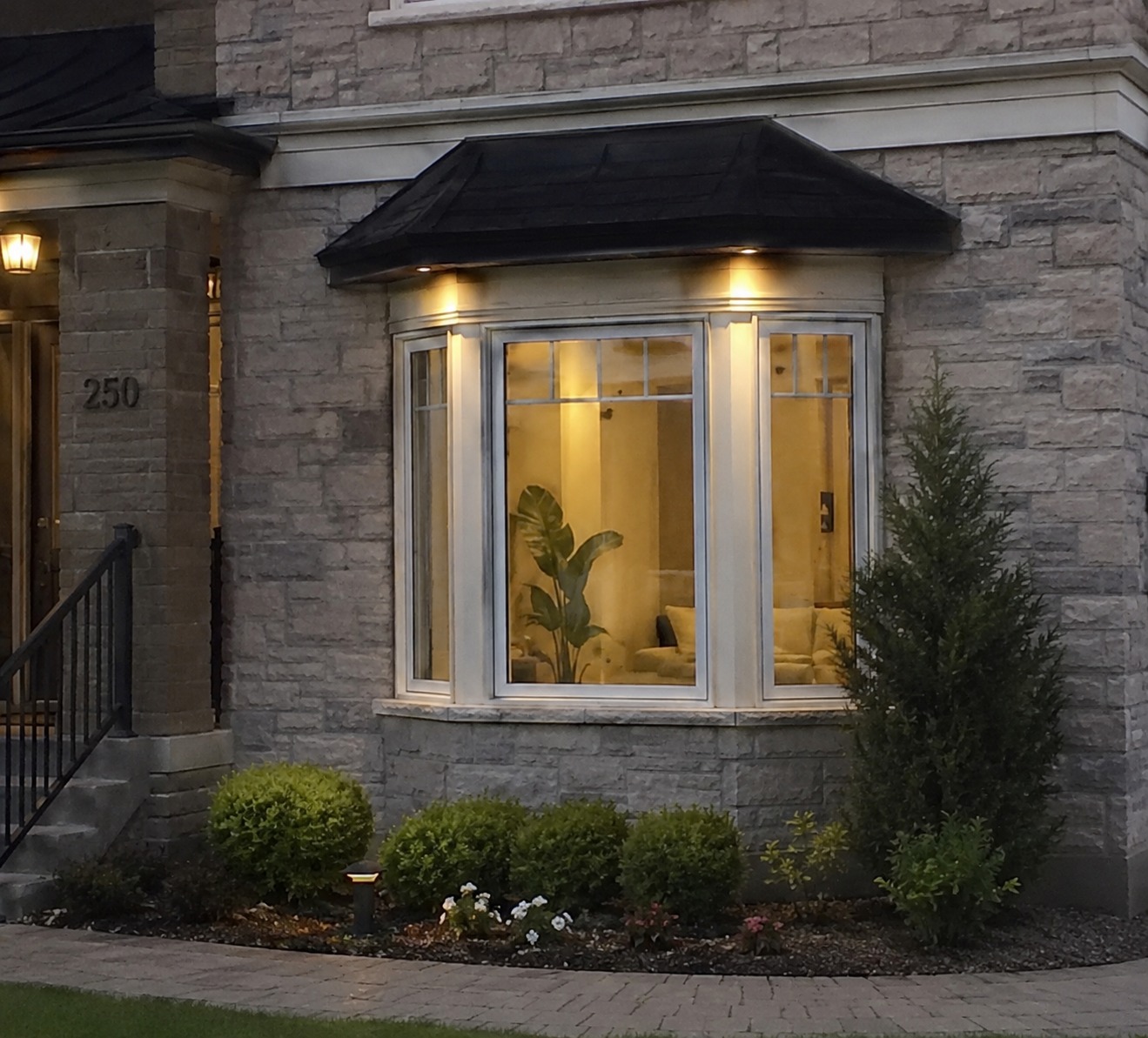 Bay window with a peaked black awning roof on a stone home exterior, lit from inside at dusk